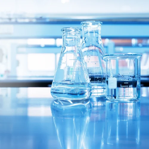 Laboratory glassware filled with clear water on a reflective blue lab bench, with lab equipment blurred in the background.